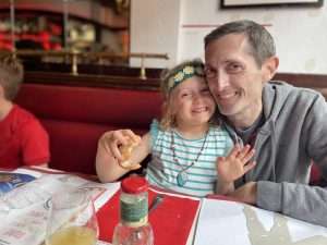 A young girl and her dad in a restaurant, he has his arm around her and they are smiling at the camera. She has a piece of bread in her hand, a striped top and a hair band with felt flowers on.
