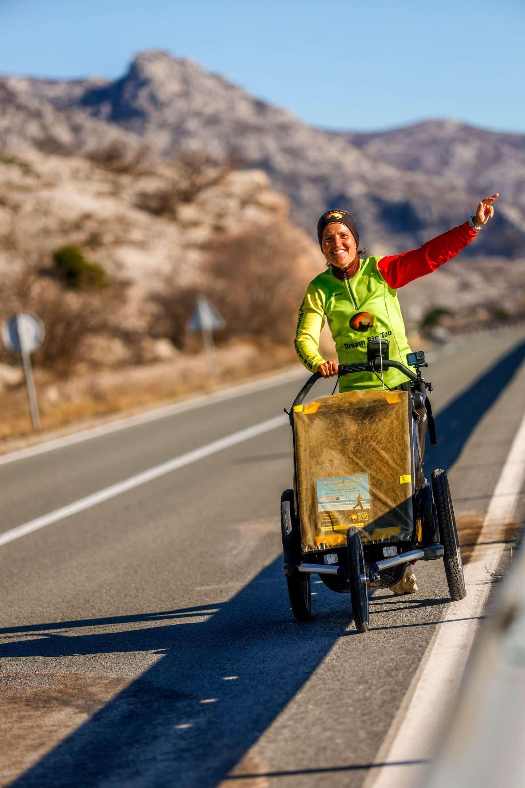 A photograph of Andrea running whilst pushing a wagon in front of mountains. Andrea is wearing sunglasses and a cap, plus running gear.
